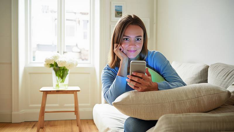 junge Frau schaut auf ihr Smartphone und liegt gemütlich auf dem Sofa